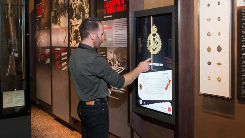 A man reaches out to touch a vertical 42 inch touchscreen with a large regimental badge on it